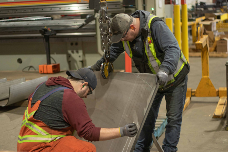 Two welders at Mainland Machinery help stabilize a piece of steel in the shop with the help of an overhead crane.
